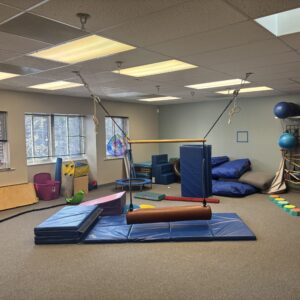 A pediatric sensory integration gym featuring a suspended bolster swing, blue crash mats, foam climbing blocks, a mini trampoline, and colorful balance stepping stones.