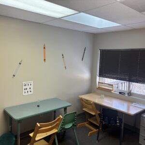 A pediatric therapy room with two worktables, ergonomic wooden and plastic chairs, a hand exercise chart, and decorative crayon wall decals.