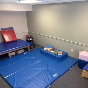 "A pediatric therapy room featuring blue floor mats, an elevated therapy table, and a variety of colorful developmental toys including a musical keyboard and stacking rings