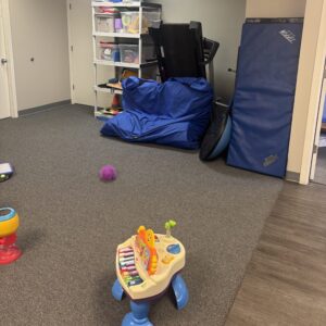 A pediatric therapy workspace featuring large blue floor mats, an elevated therapy table, a red incline wedge, and various developmental toys for fine motor skill practice.